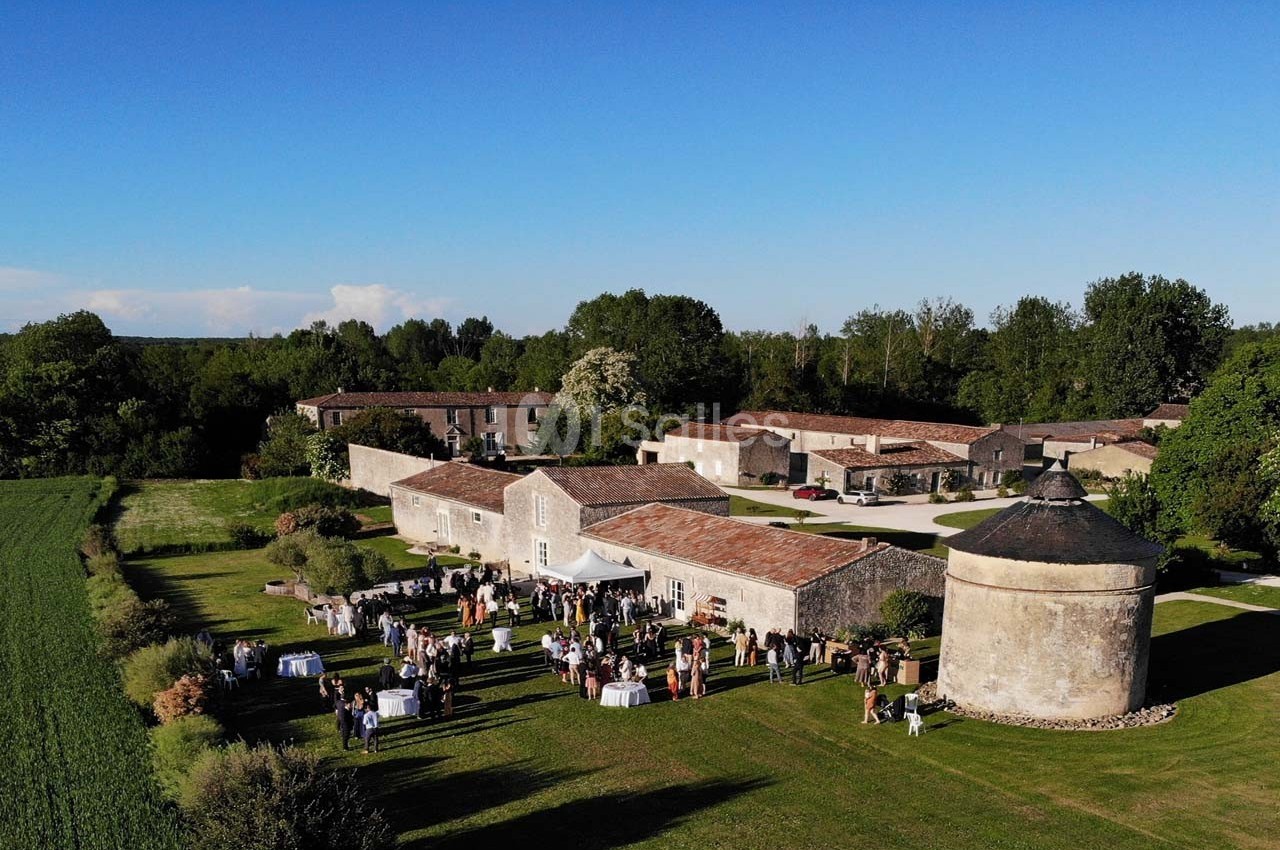 Vue aérienne d'un domaine rural avec des bâtiments en pierre, une tour ronde et un rassemblement en extérieur.