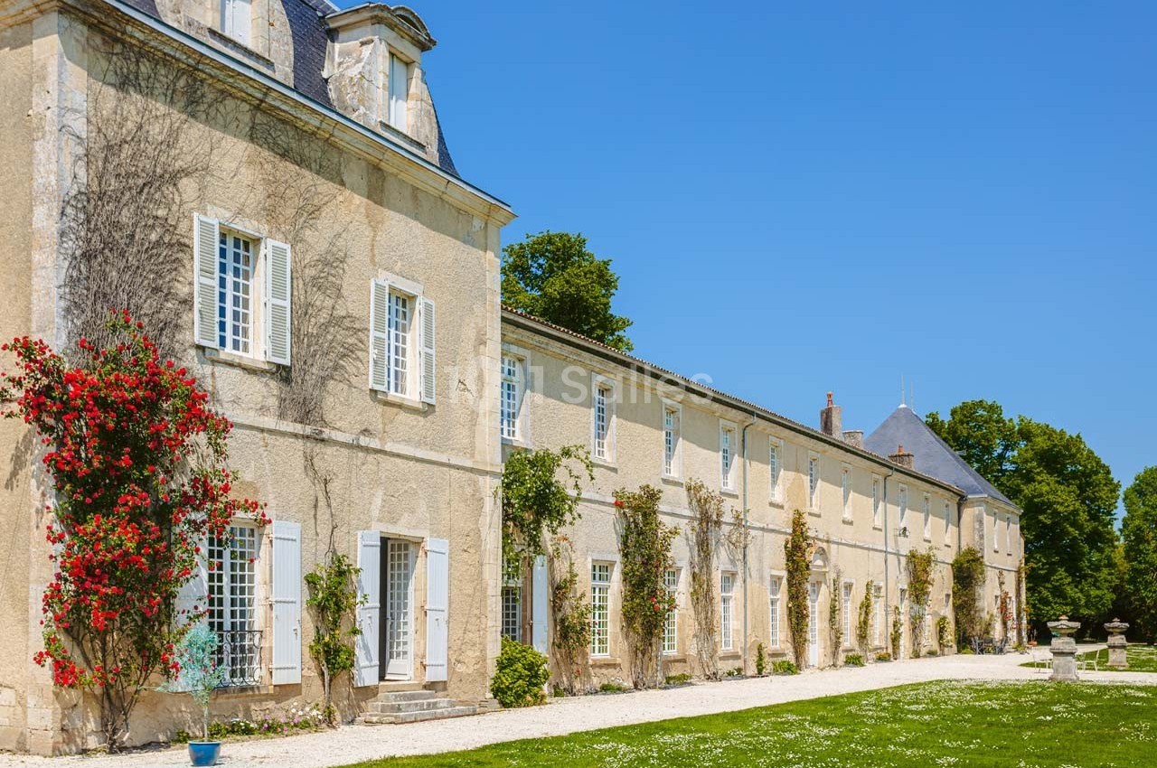 Façade d'un grand bâtiment en pierre avec volets blancs, entouré de végétation et d'un jardin sous un ciel bleu.