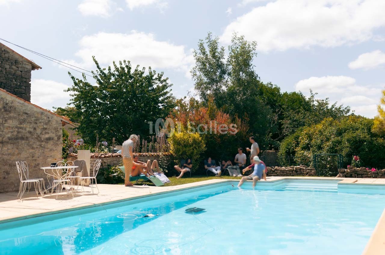 Personnes profitant d'une piscine extérieure dans un jardin ensoleillé avec des arbres et des buissons en arrière-plan.