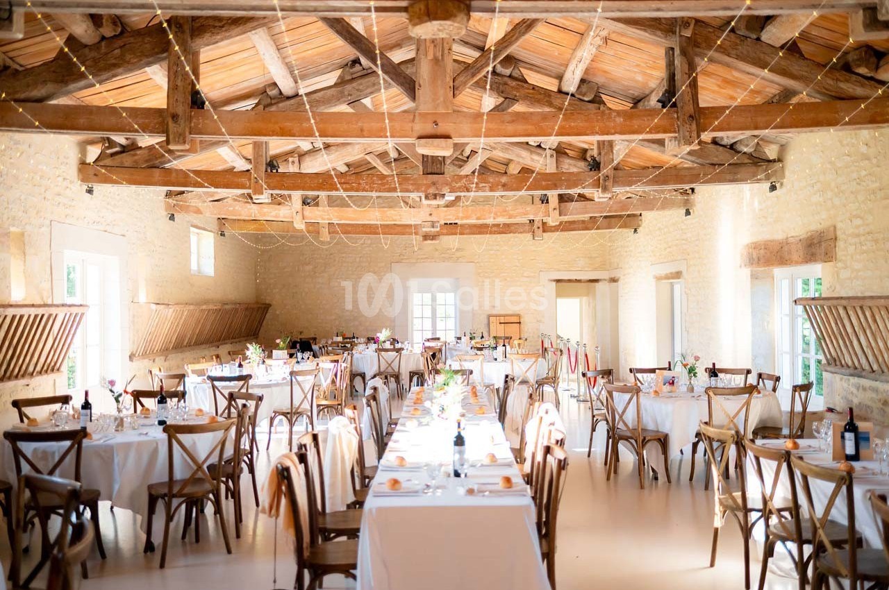 Salle de réception lumineuse avec tables dressées, chaises en bois et poutres apparentes au plafond.