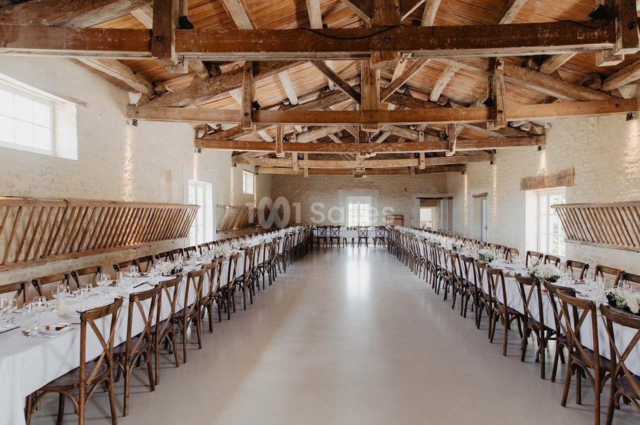 Salle de réception lumineuse avec poutres apparentes, tables longues dressées et chaises en bois alignées.