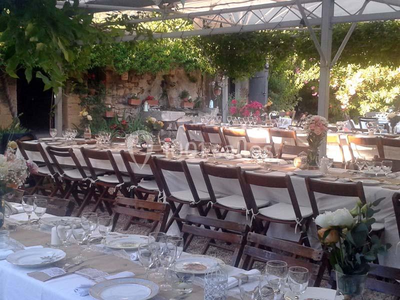 Tables dressées pour un repas en extérieur sous une pergola, entourées de verdure et de fleurs.