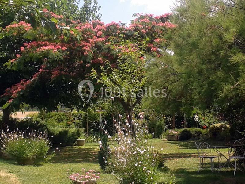 Jardin verdoyant avec des arbres fleuris, des buissons et un mobilier de jardin en métal blanc sur une pelouse.
