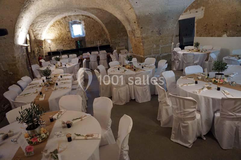 Salle voûtée en pierre aménagée pour un événement, avec des tables rondes décorées et des chaises couvertes de housses…