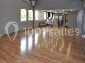 Salle vide avec parquet en bois, murs gris, grandes fenêtres laissant entrer la lumière naturelle et un coin salon.