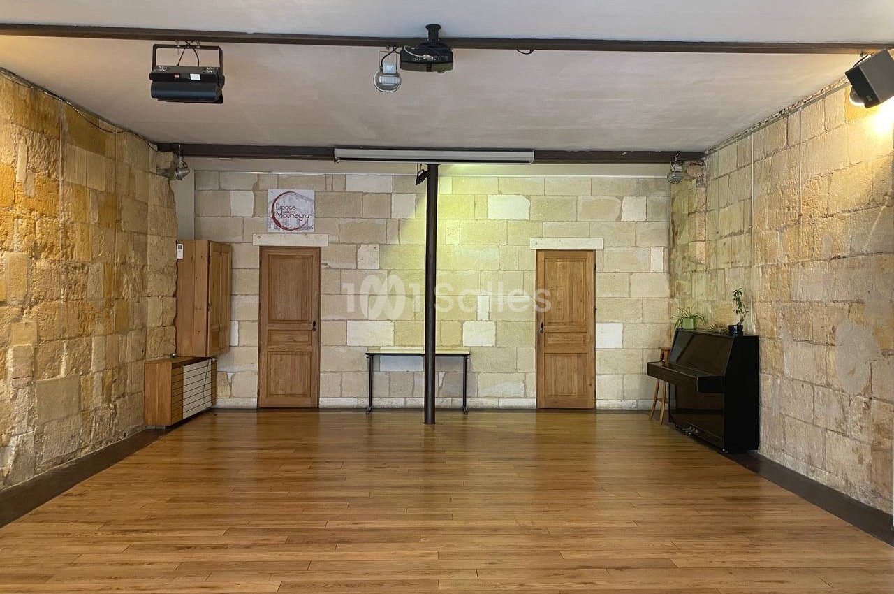 Salle vide avec parquet en bois, murs en pierre, trois portes en bois, un piano noir et des projecteurs au plafond.