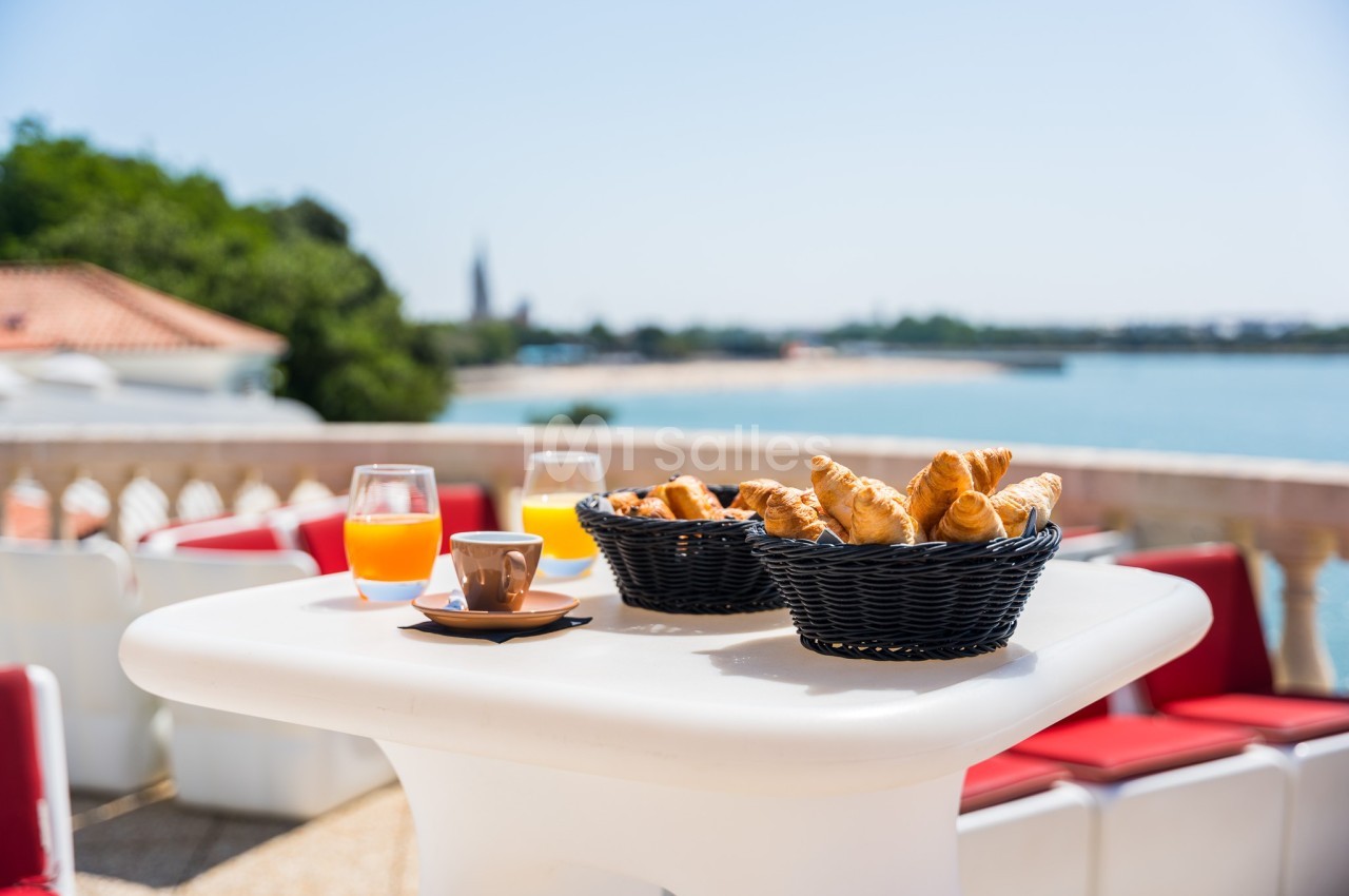 Petit-déjeuner avec croissants, jus d'orange et café sur une table en terrasse avec vue sur un paysage côtier.