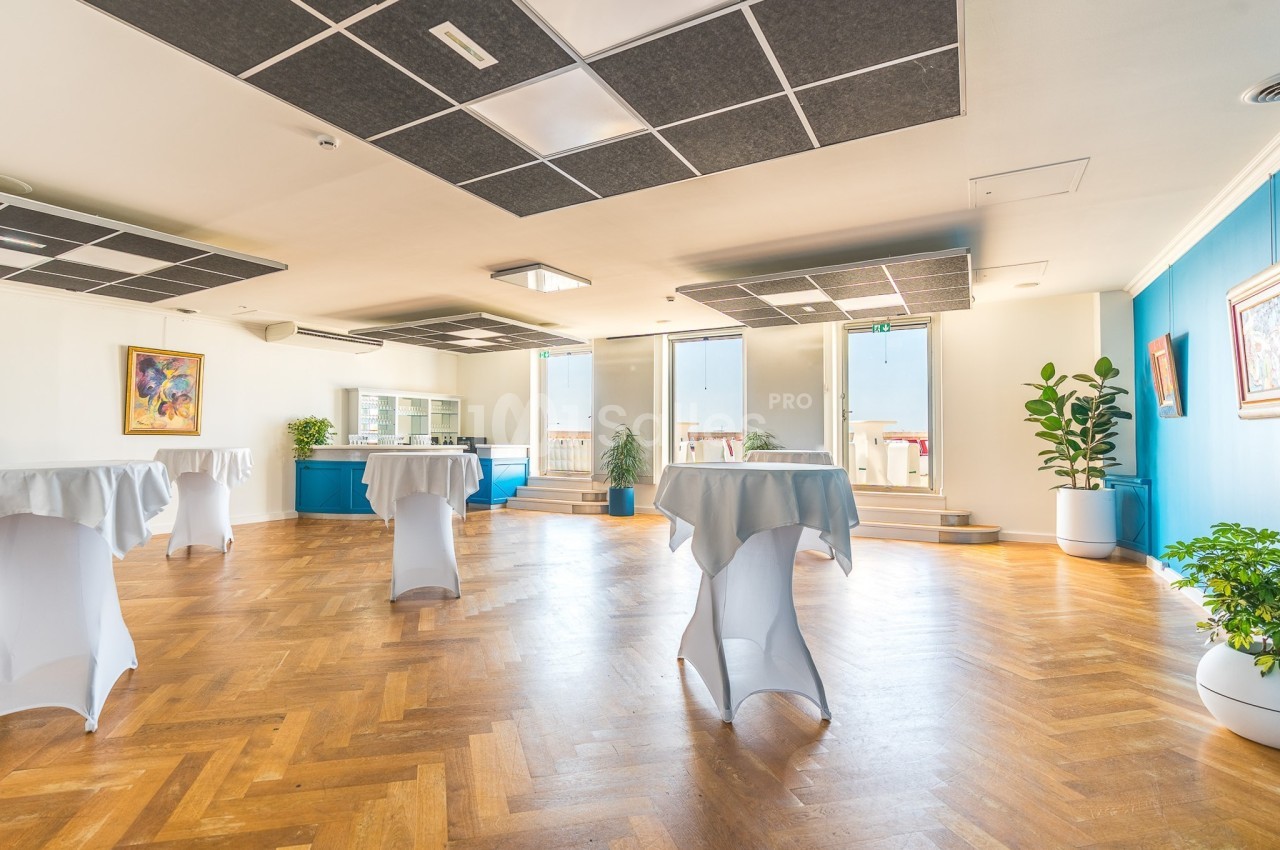 Salle lumineuse avec parquet en bois, tables hautes couvertes de nappes blanches et plantes décoratives.