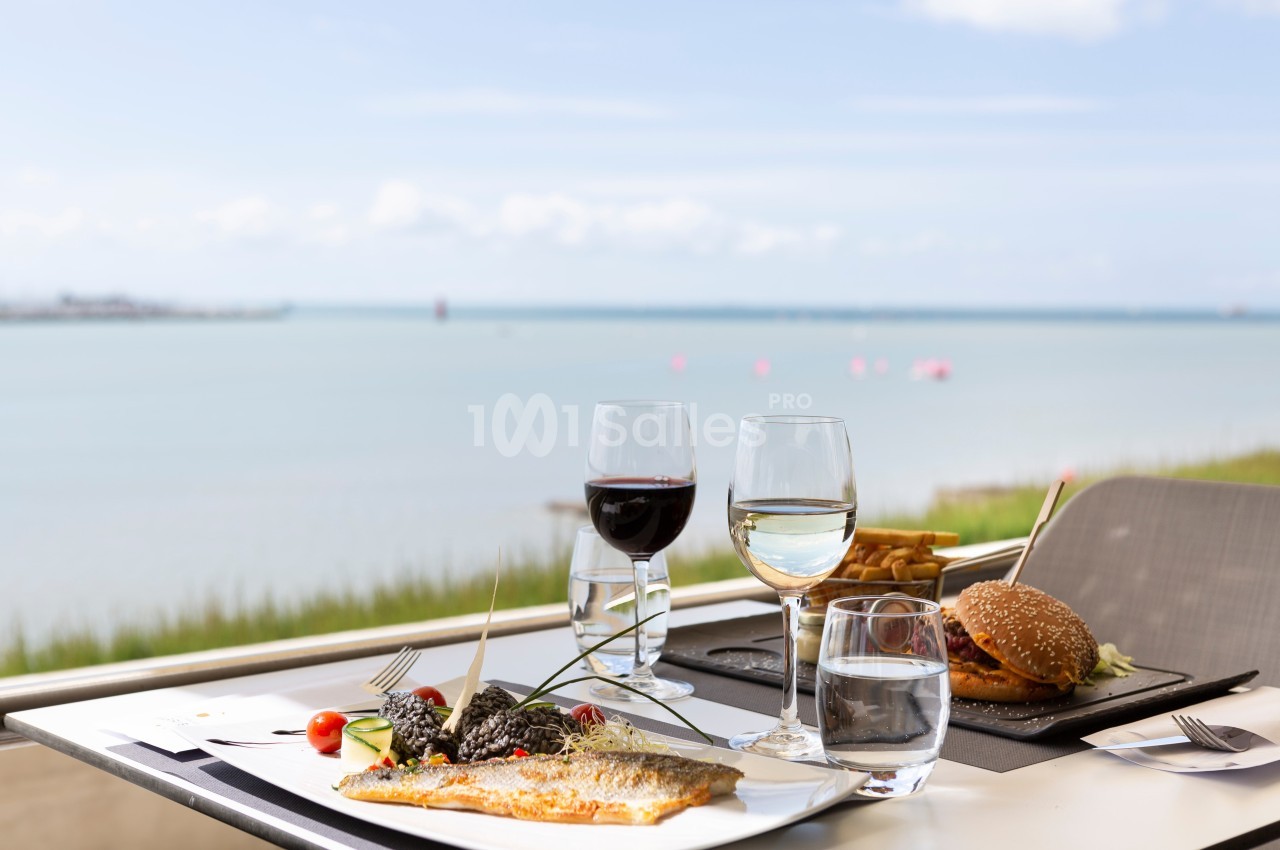 Repas avec poisson, burger, vin et eau servis sur une table en terrasse avec vue sur la mer.