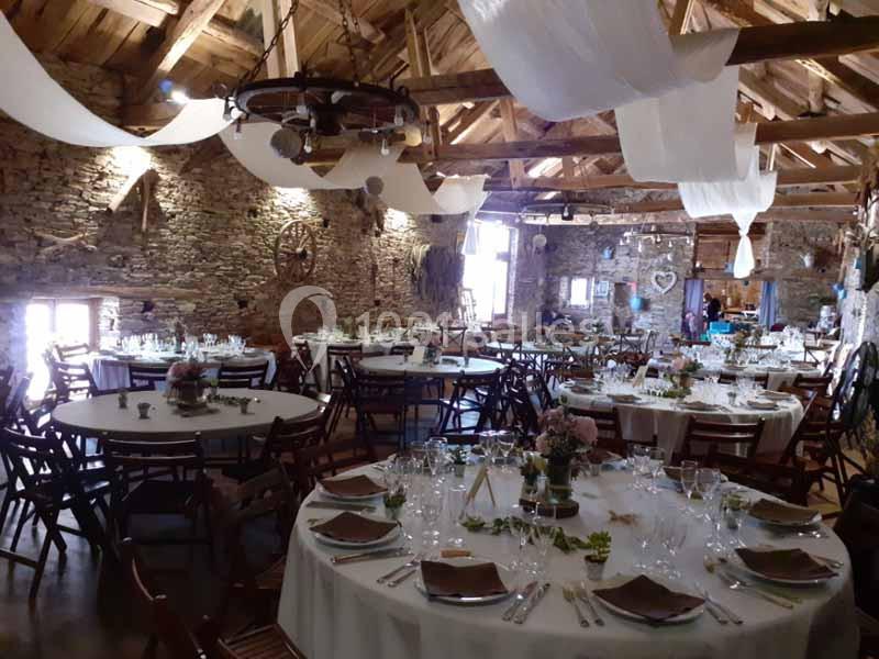 Salle de réception rustique avec tables rondes dressées, poutres apparentes et murs en pierre.