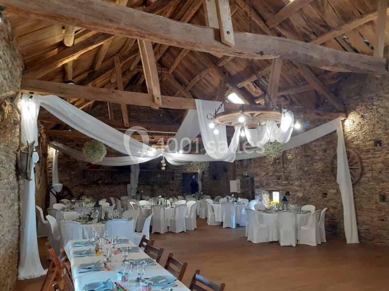 Salle rustique avec murs en pierre, poutres apparentes et tables dressées pour un événement, décorée de voilages blancs.