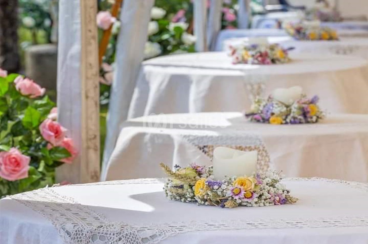 Tables décorées de nappes blanches, ornées de bougies entourées de fleurs, dans un cadre extérieur fleuri.
