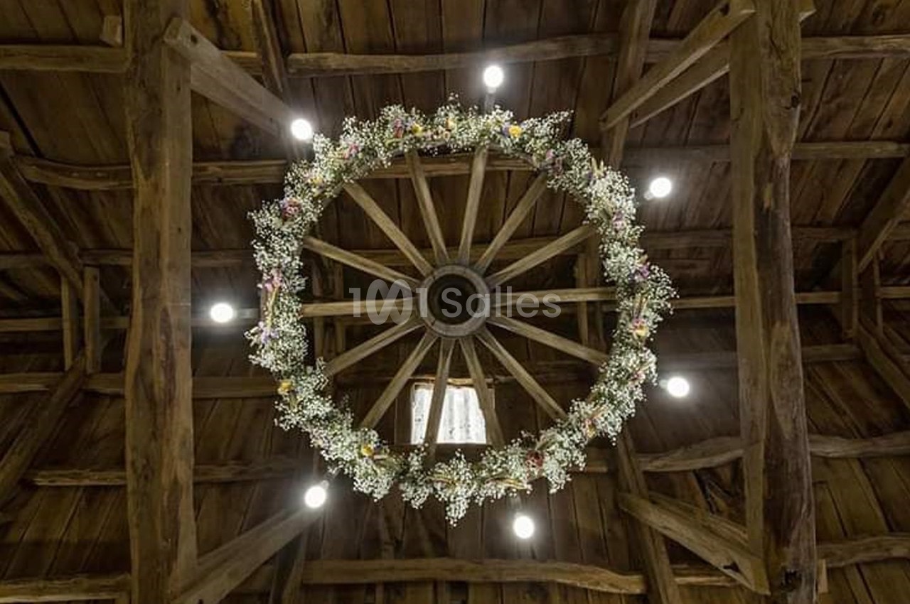 Vue d'une charpente en bois avec une roue décorée de fleurs suspendue au plafond, éclairée par des spots lumineux.