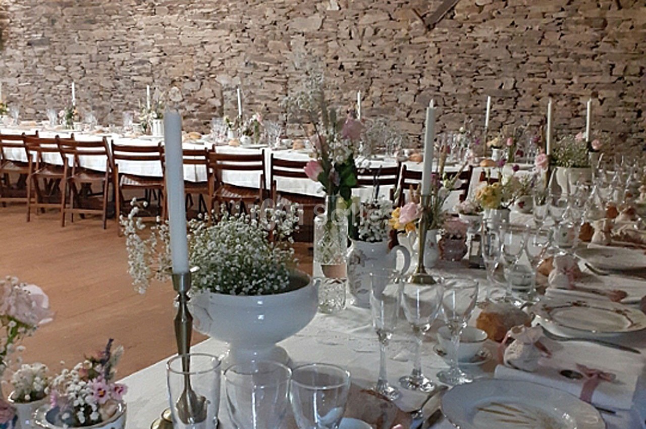 Grande table de réception décorée avec des fleurs, bougies et vaisselle, dans une salle aux murs en pierre apparente.