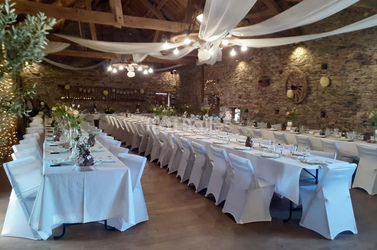 Salle de réception décorée avec des tables alignées, nappes blanches et guirlandes lumineuses sous un plafond en bois.