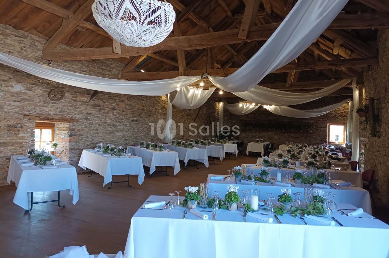 Salle de réception en pierre avec tables décorées de nappes blanches et centres de table fleuris, sous des drapés suspendus.