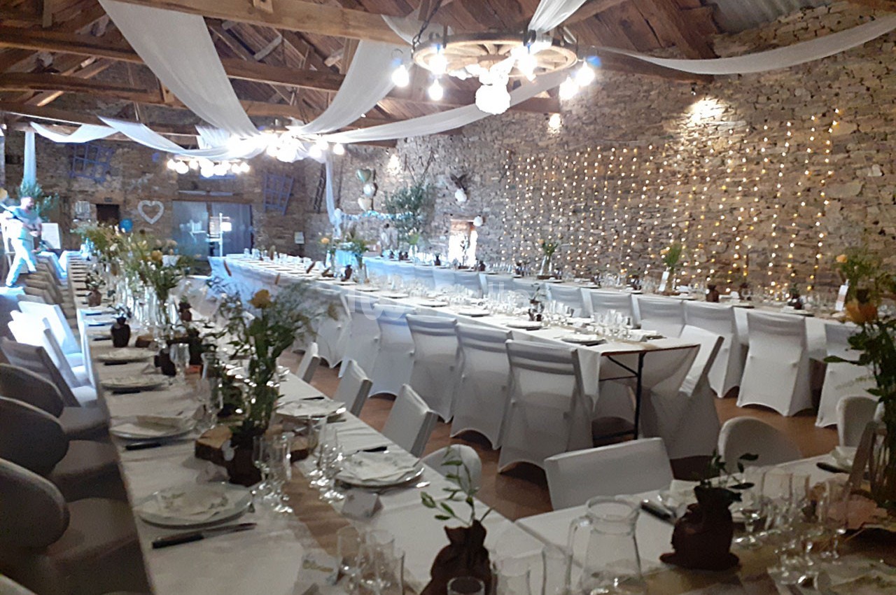 Grande salle de réception décorée pour un événement, avec longues tables dressées, chaises blanches et guirlandes lumineuses.