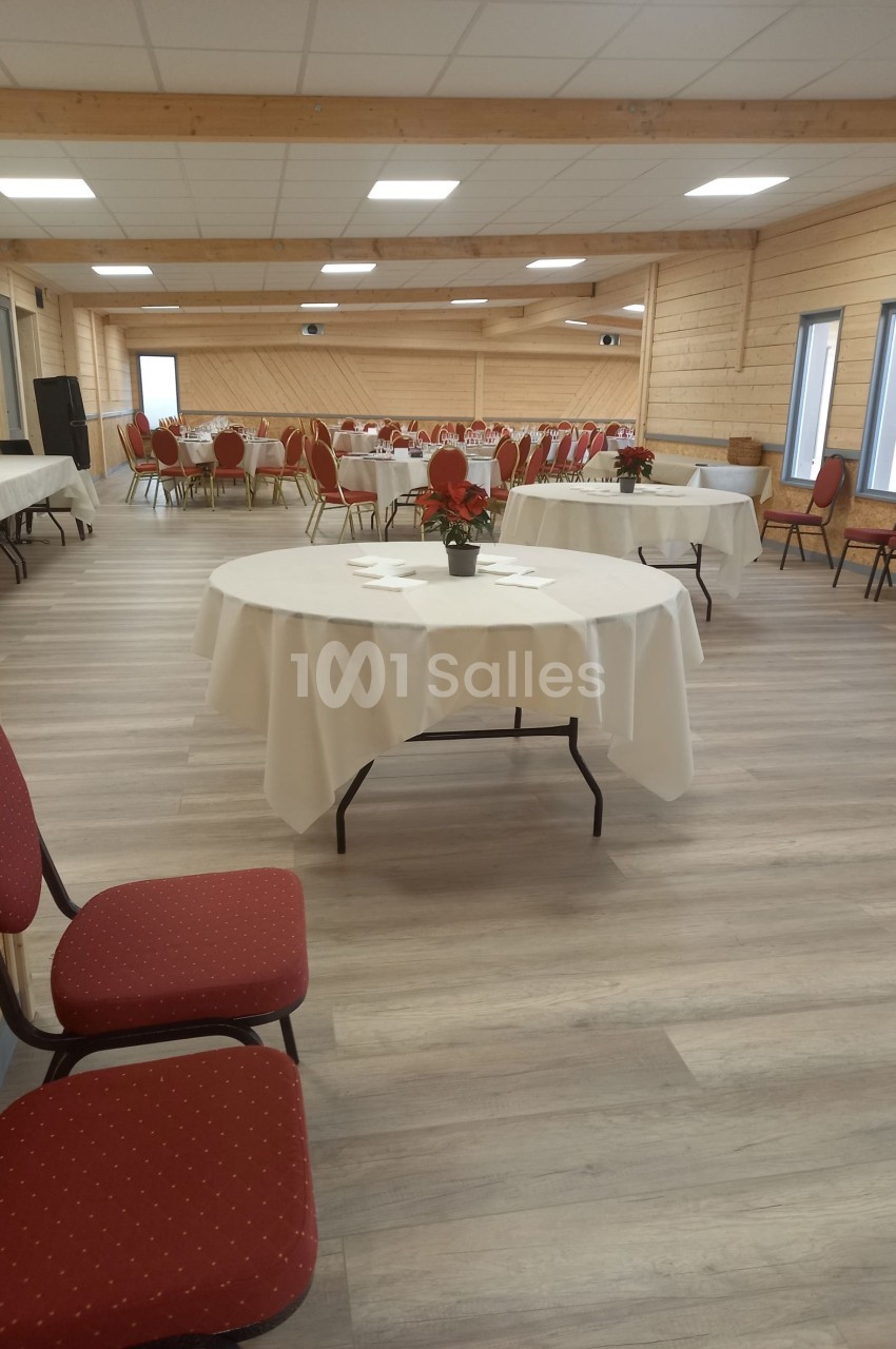 Salle de réception lumineuse avec tables rondes dressées, nappes blanches et chaises rouges.