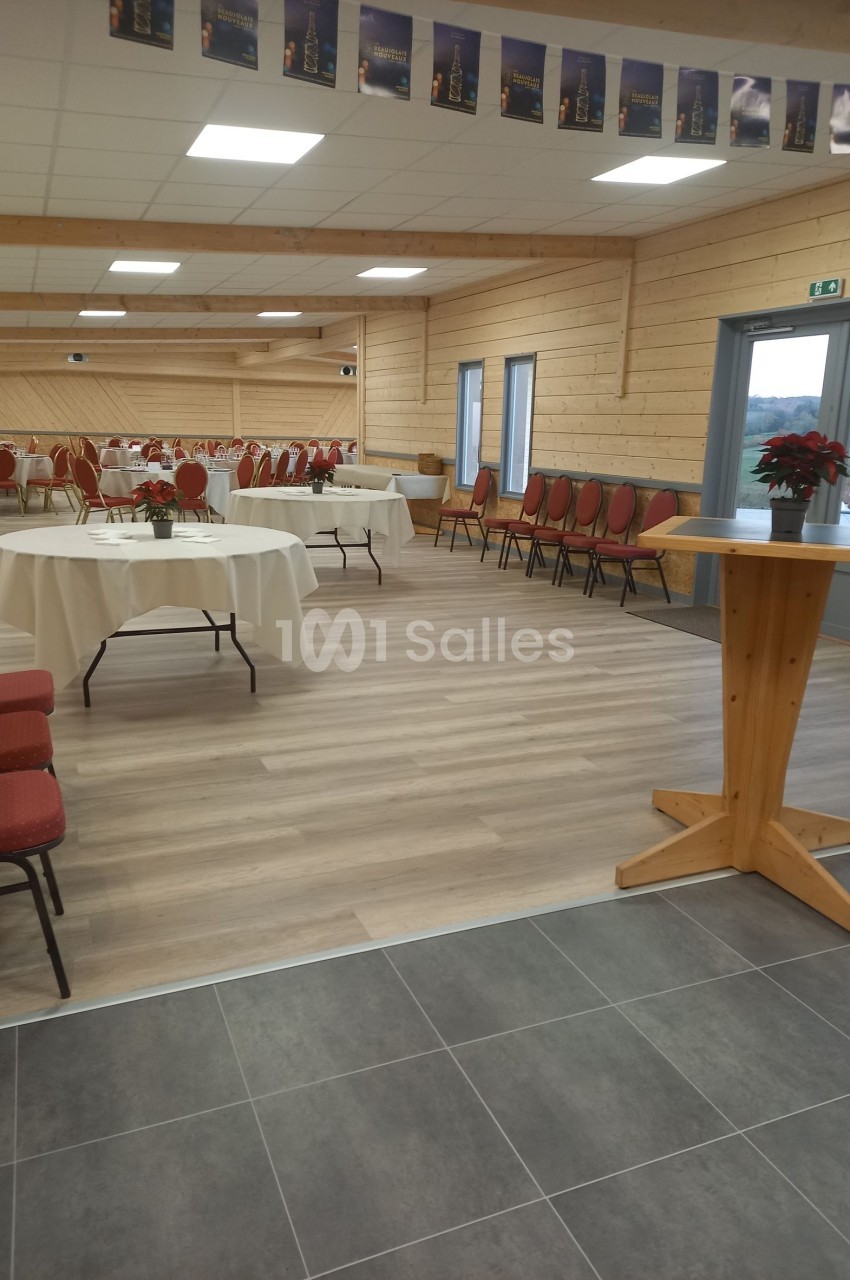 Salle de réception lumineuse avec tables rondes dressées, chaises rouges et décorations simples.