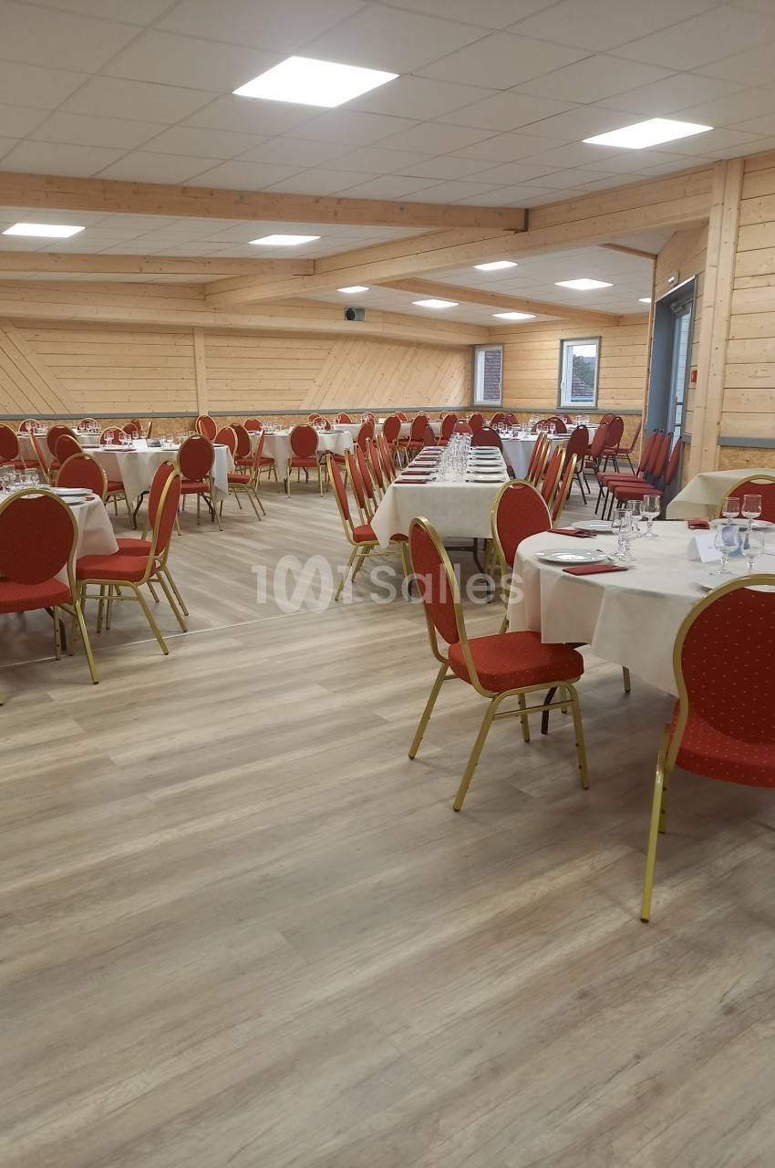 Salle de réception avec tables rondes dressées, chaises rouges et murs en bois clair.