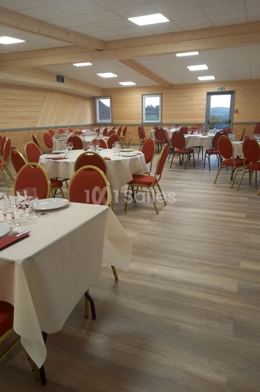 Salle de réception lumineuse avec tables dressées, nappes blanches et chaises rouges, sol en parquet clair.