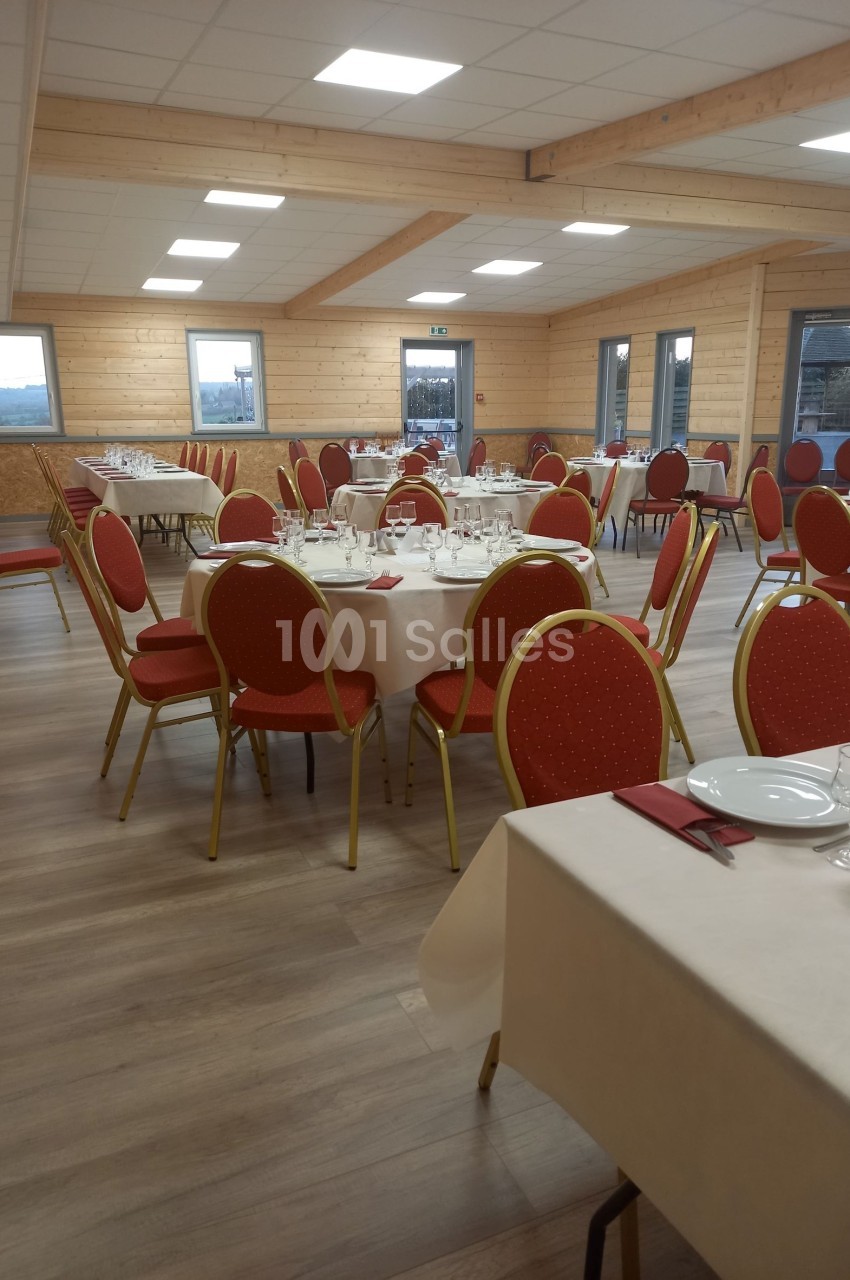 Salle de réception avec tables rondes dressées, nappes blanches, serviettes rouges et chaises rouges sur un sol en bois.