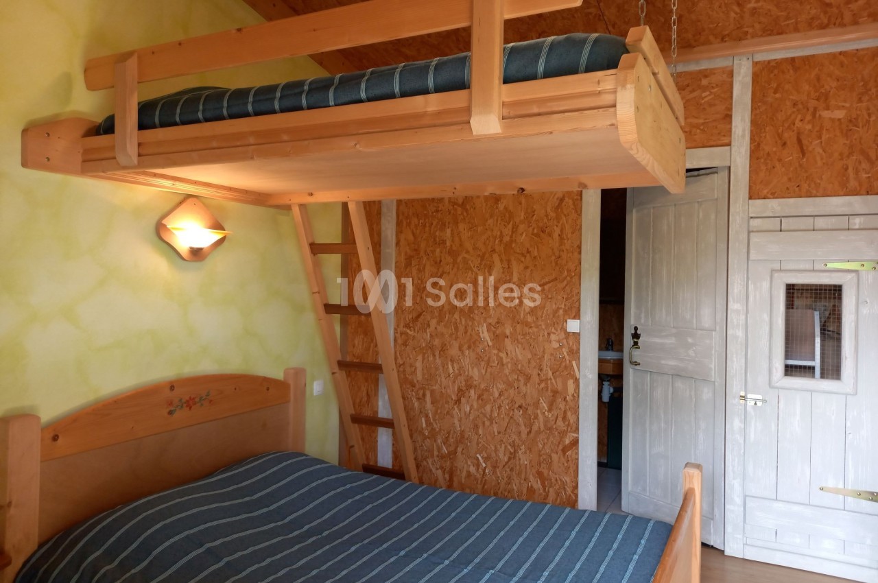 Chambre en bois avec lit double, mezzanine avec matelas, échelle en bois et porte blanche sur un mur texturé.
