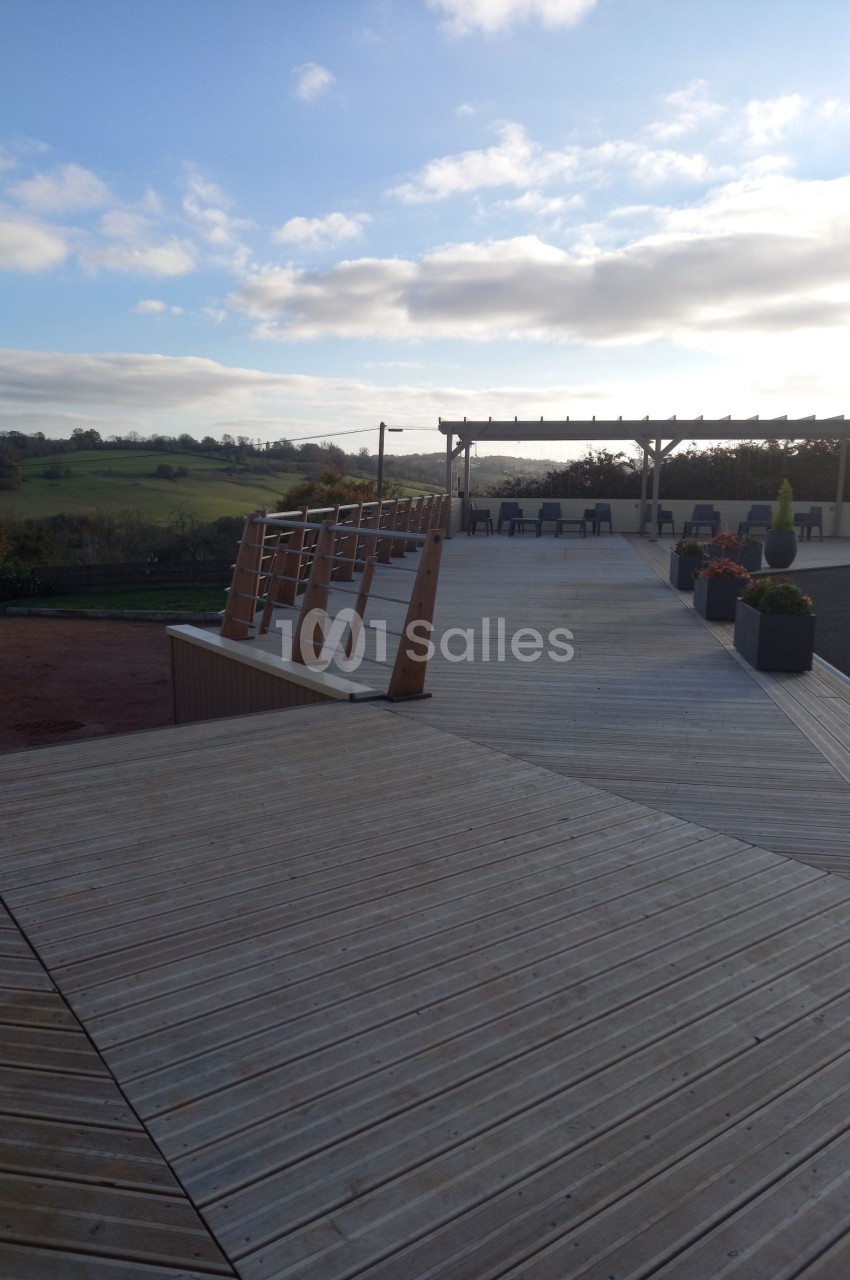Terrasse en bois avec rampe d'accès, pergola et vue dégagée sur un paysage rural sous un ciel partiellement nuageux.