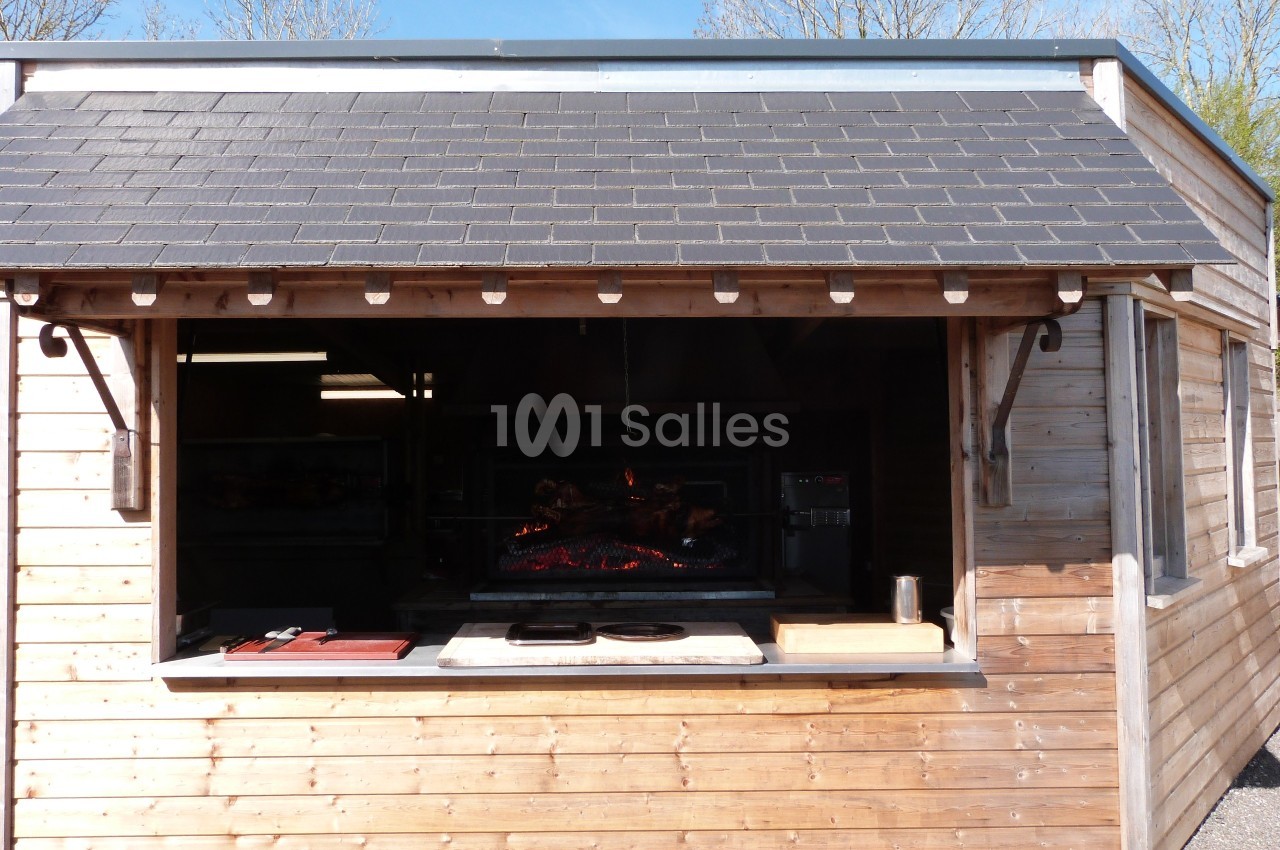 Abri en bois avec un four à rôtisserie visible à travers une ouverture, entouré d'arbres sous un ciel dégagé.