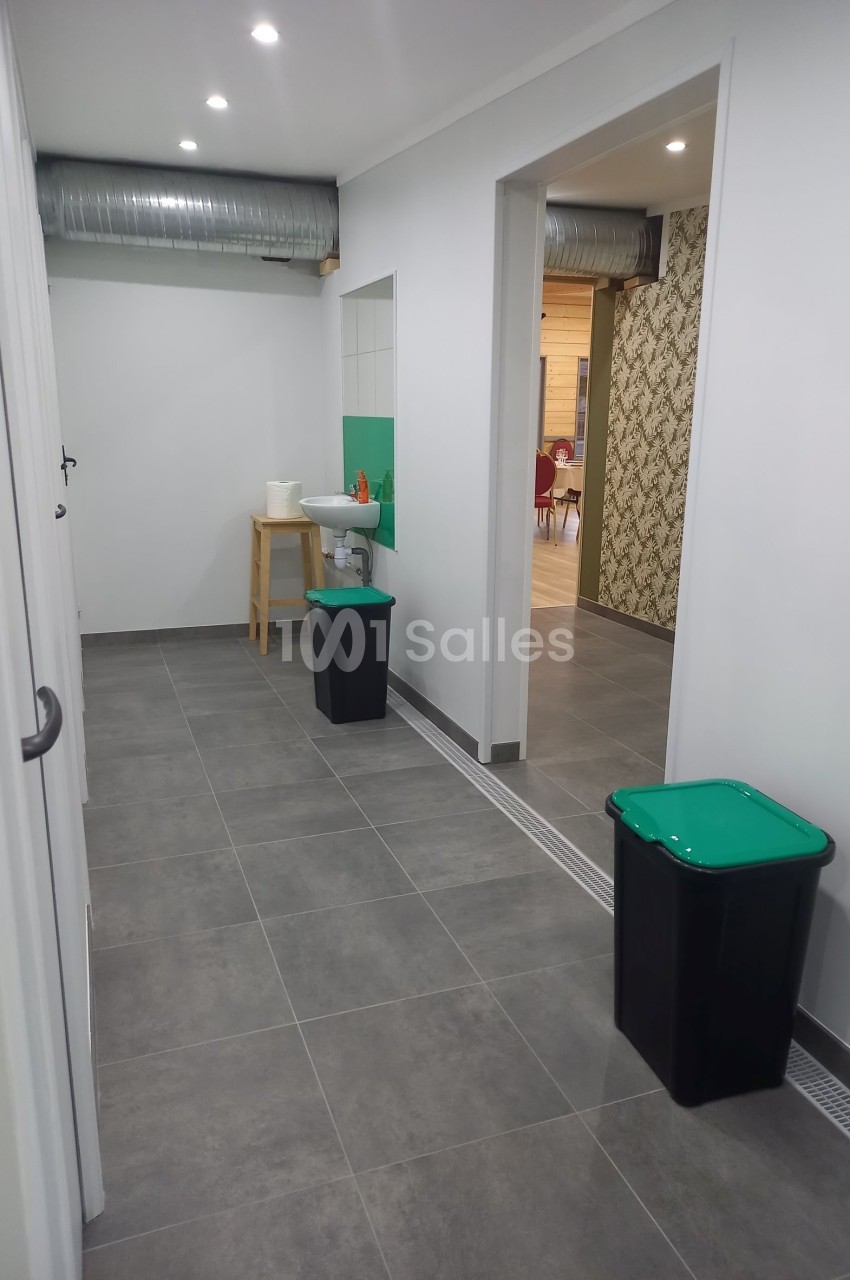 Couloir avec sol en carrelage gris, deux poubelles à couvercle vert, un lavabo et un mur décoré en arrière-plan.