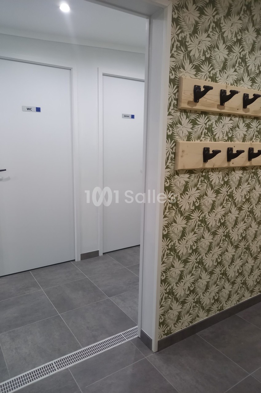 Vue d'un couloir avec des portes blanches de toilettes et un mur décoré de papier peint à motifs végétaux, équipé de patères.