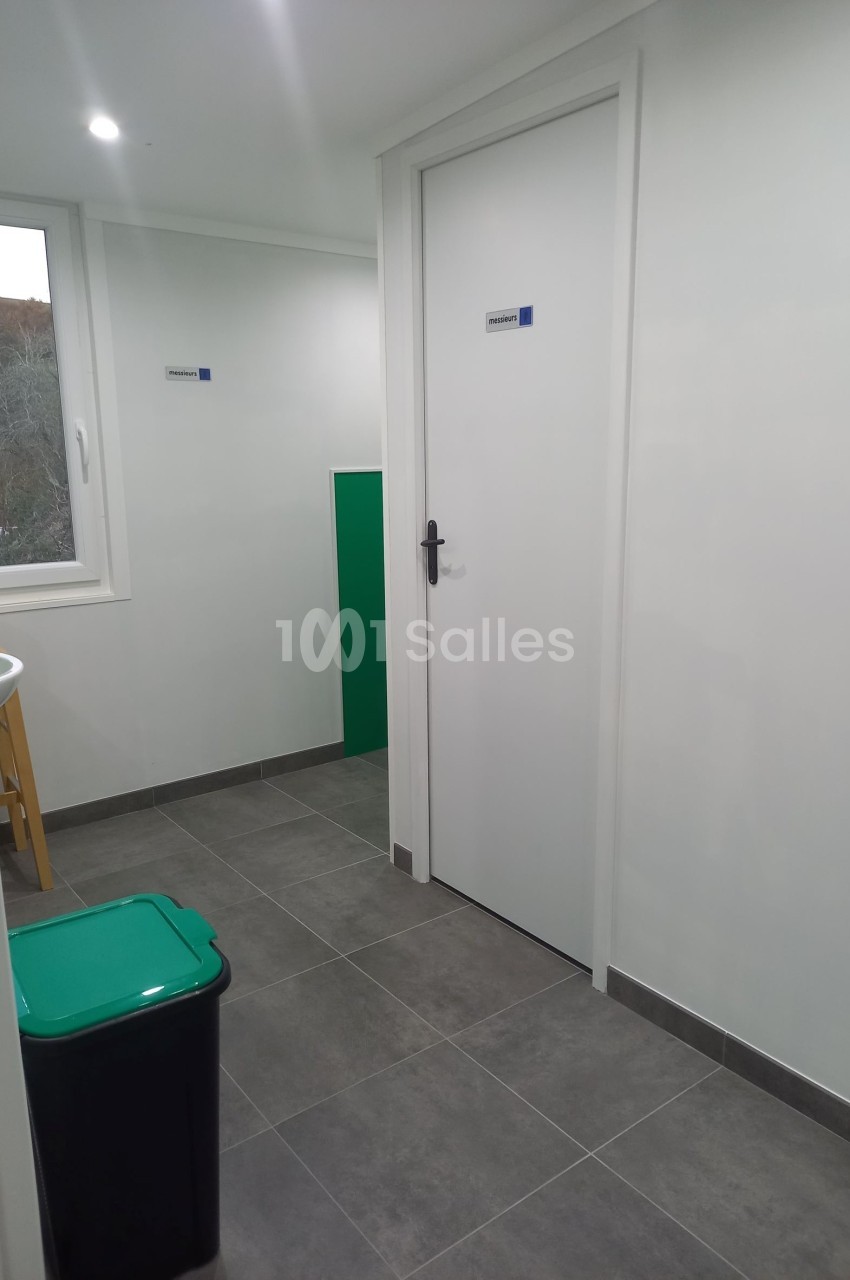 Vue d'un couloir avec sol en carrelage gris, une porte blanche fermée, une poubelle verte et une fenêtre.