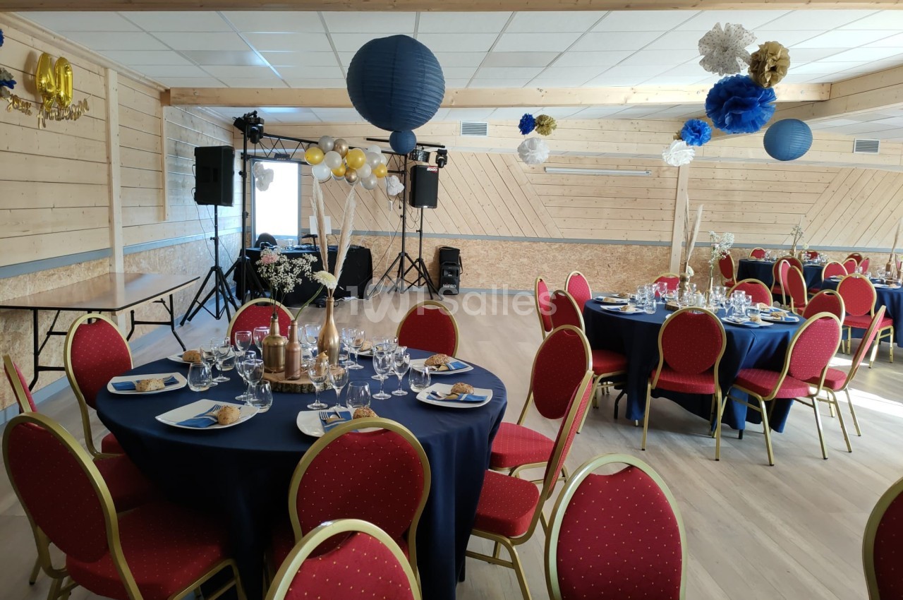 Salle décorée pour un événement avec tables rondes, nappes bleues, chaises rouges et suspensions en papier.