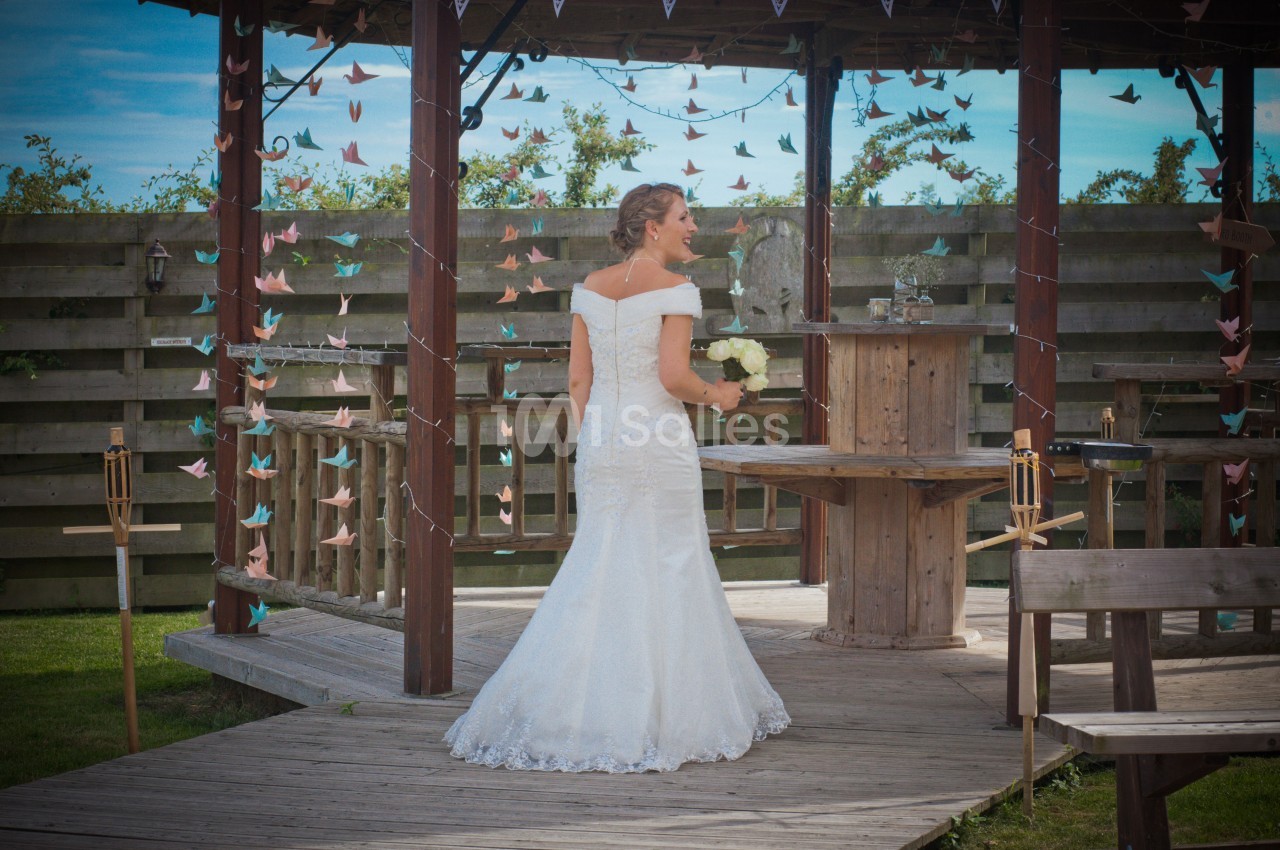 Une femme en robe de mariée blanche tient un bouquet de fleurs, debout sur une terrasse en bois décorée.