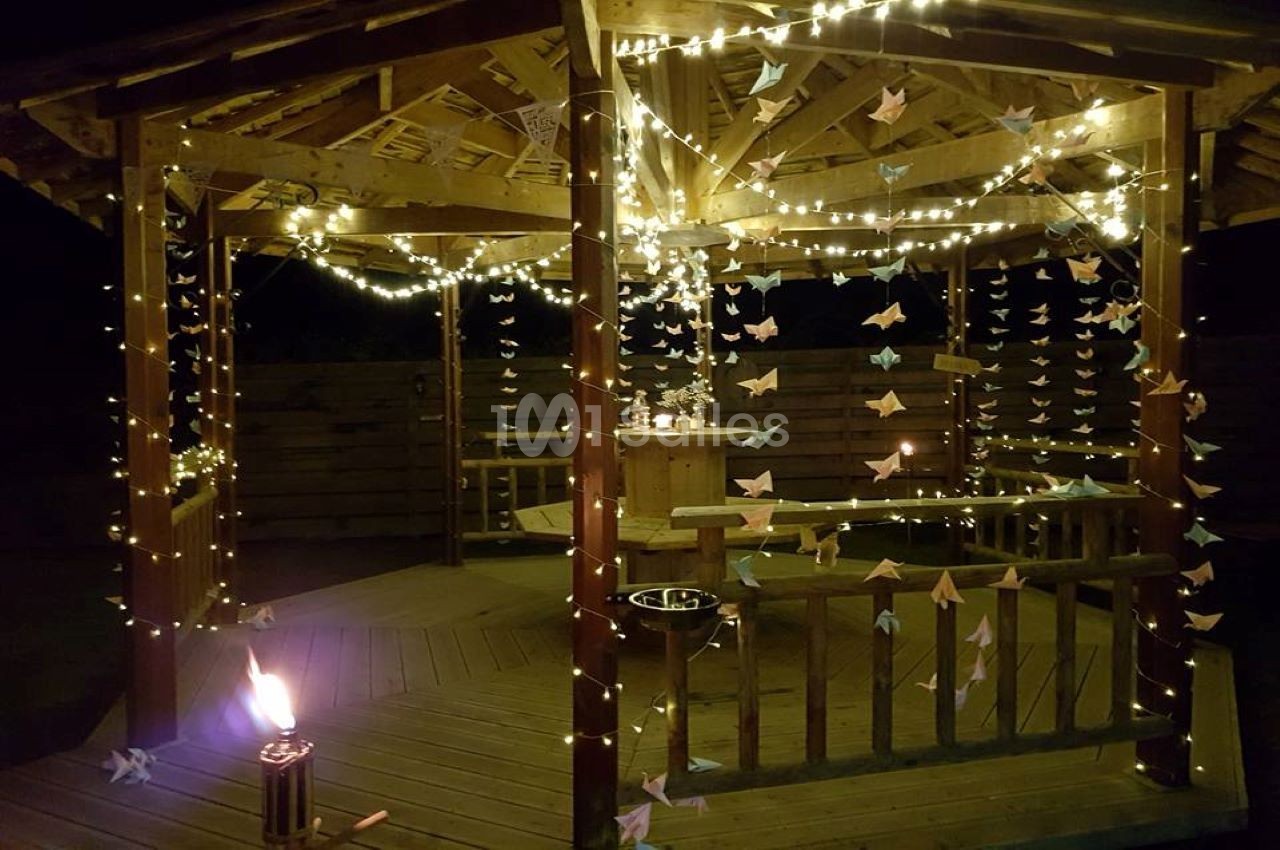 Kiosque en bois décoré de guirlandes lumineuses et d'origamis, éclairé par une torche dans une ambiance nocturne.