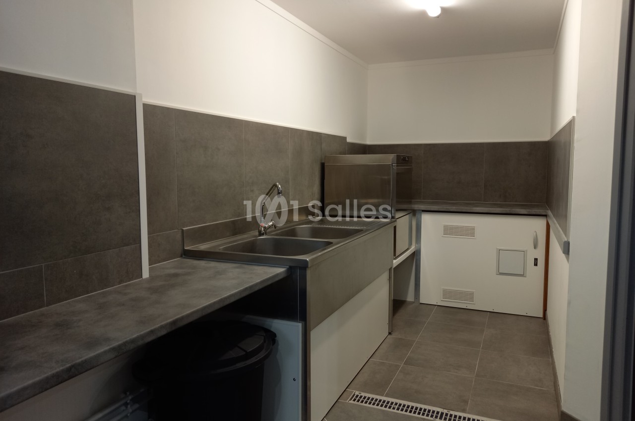 Cuisine professionnelle avec évier inox, plan de travail gris, carrelage mural et sol gris, éclairée par une lampe au…