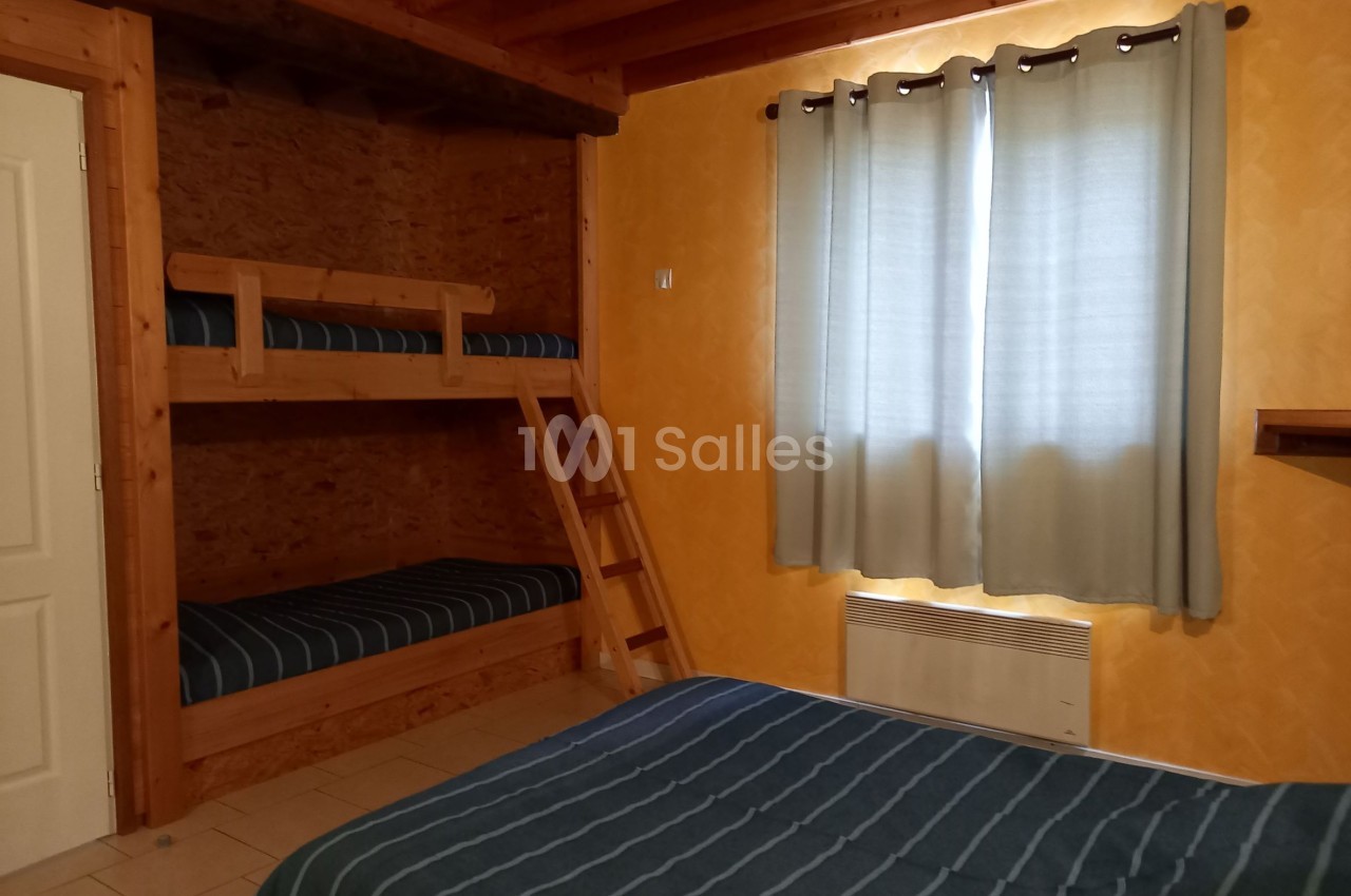 Chambre avec lits superposés en bois, lit double, murs jaunes, rideaux gris et plafond avec poutres apparentes.