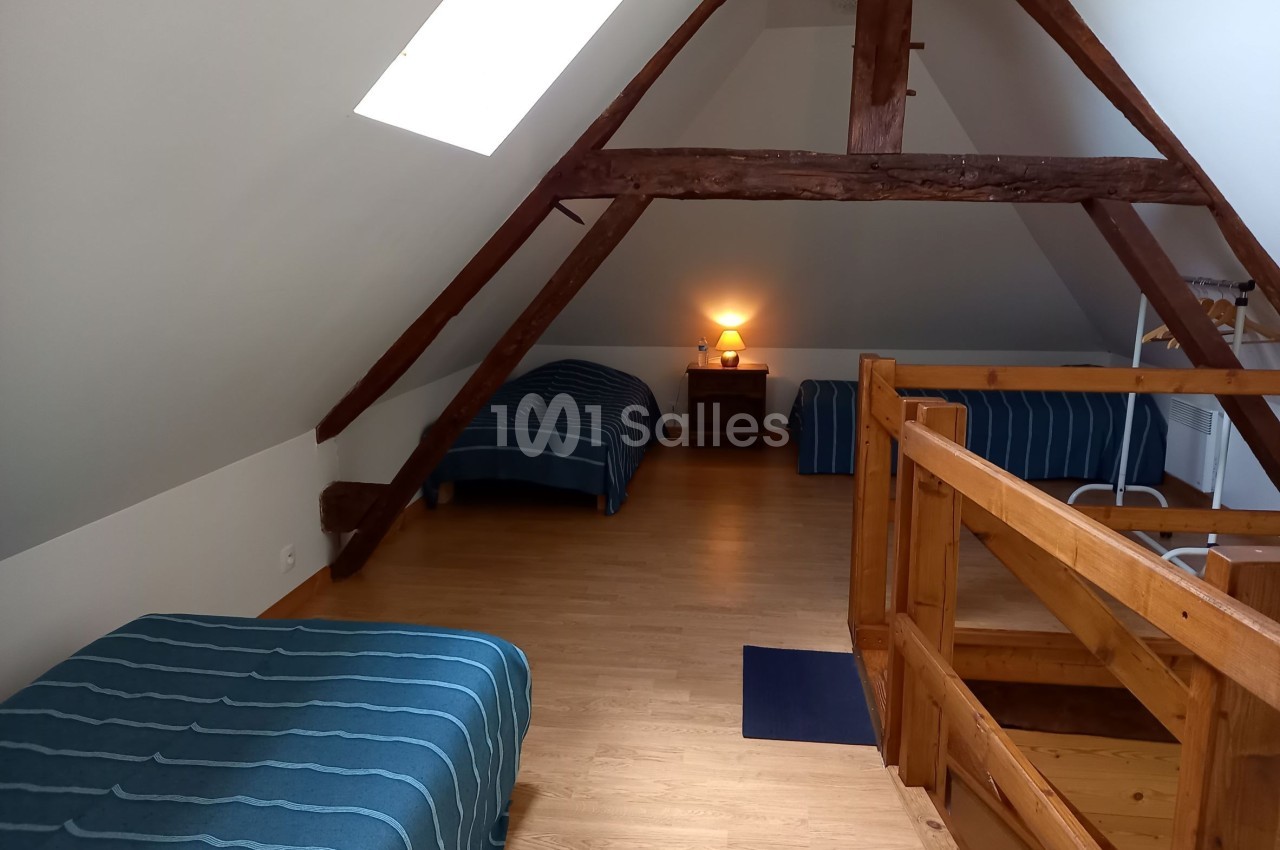 Chambre mansardée avec trois lits simples, parquet en bois, poutres apparentes et lumière douce sur une table de chevet.