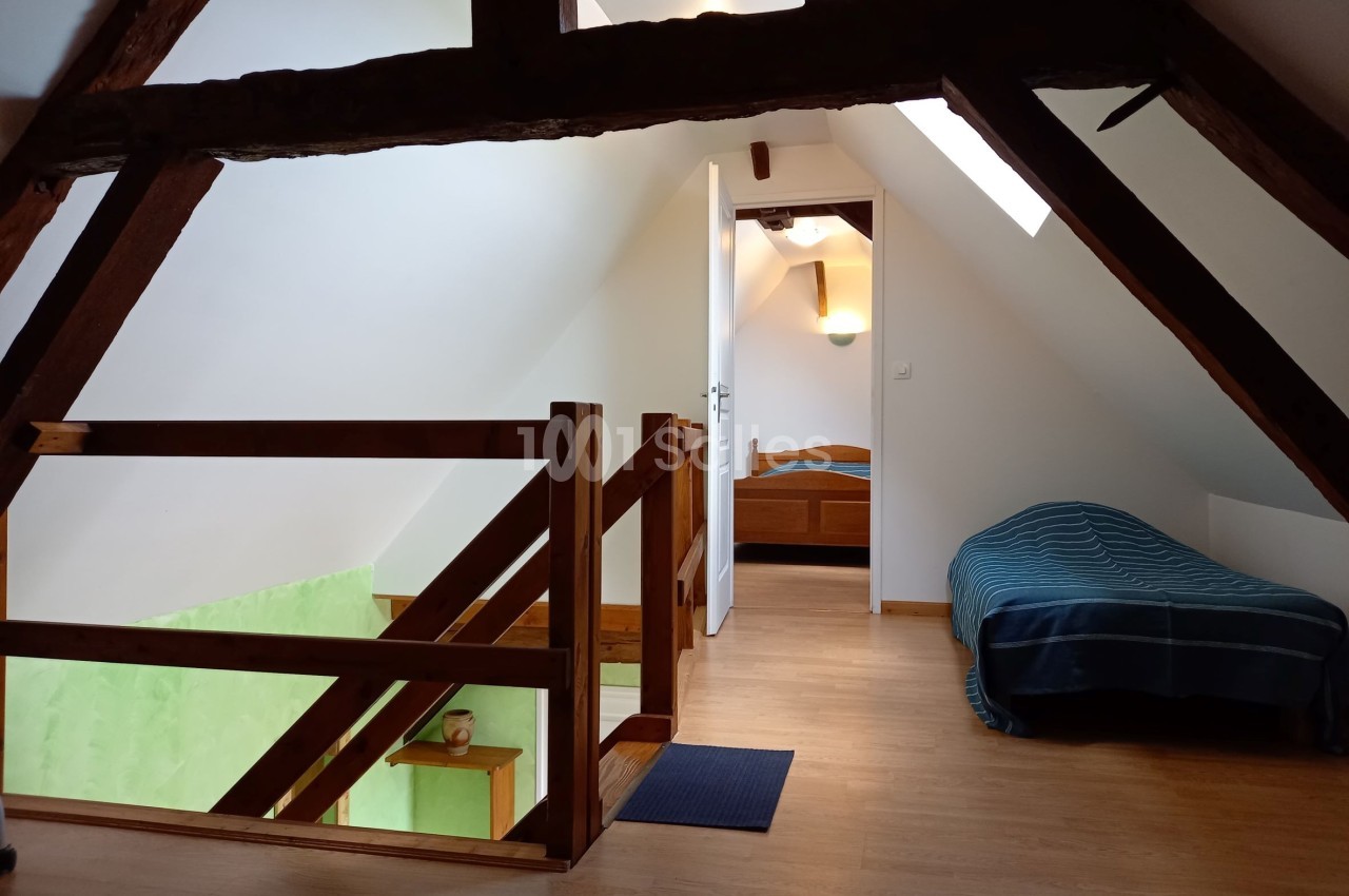 Chambre mansardée avec poutres apparentes, lit simple, lit double au fond et lumière naturelle entrant par un velux.
