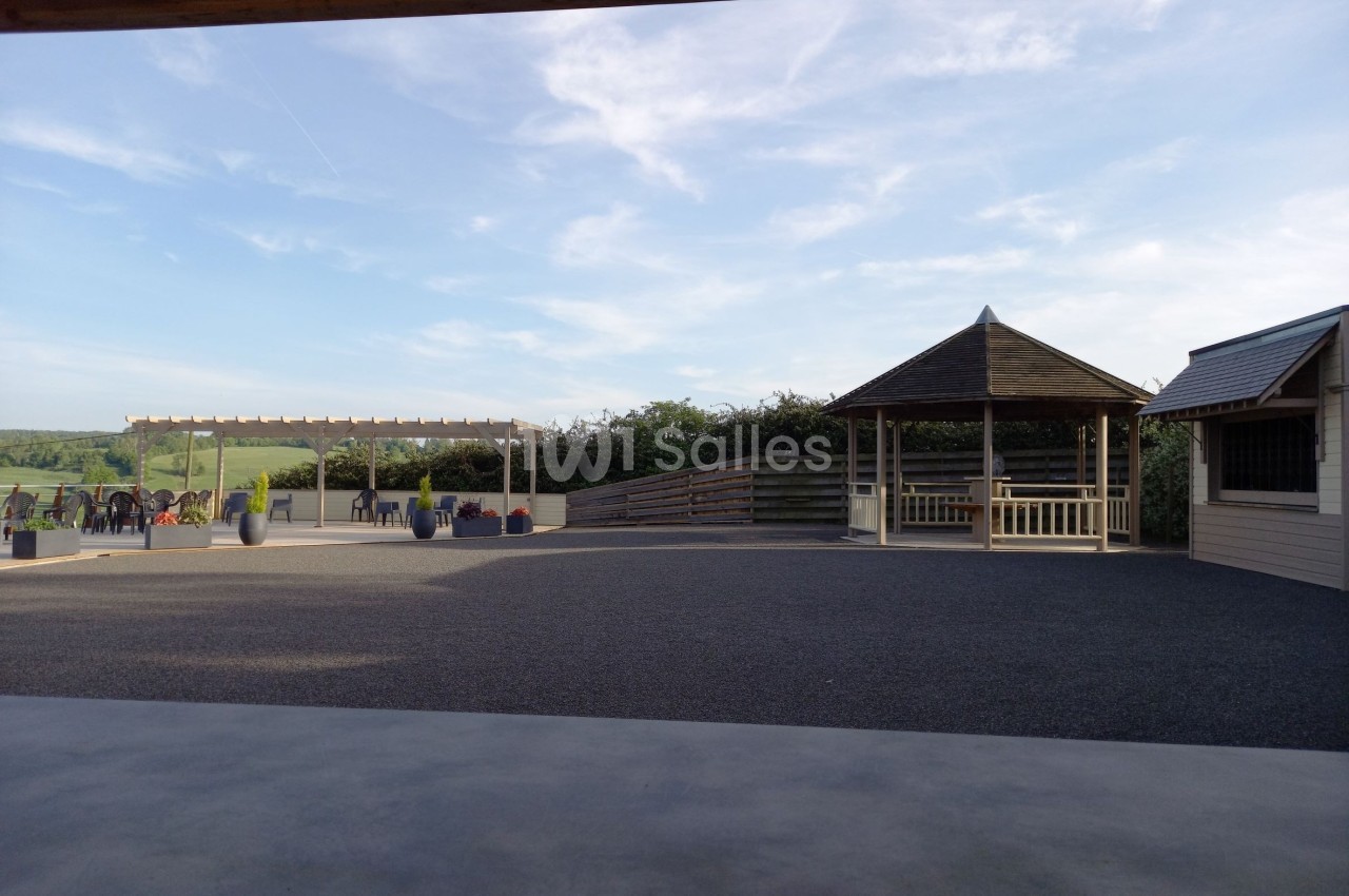 Cour extérieure avec un kiosque en bois, une pergola et des tables, entourée de verdure sous un ciel dégagé.