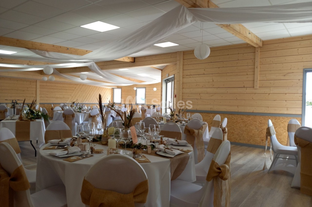 Salle de réception décorée avec des tables rondes, nappes blanches, chaises ornées de nœuds beige et voilages suspendus.