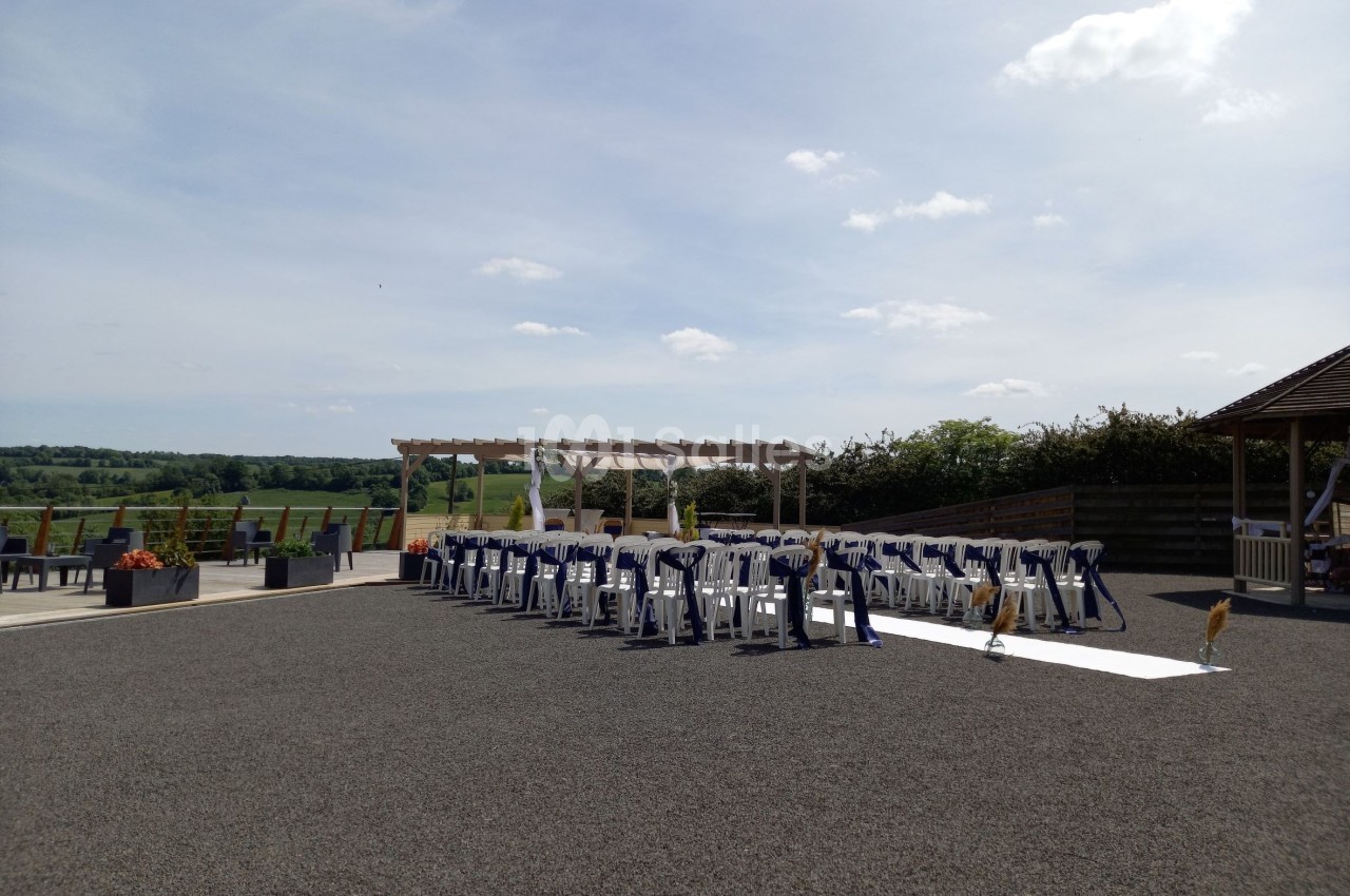 Chaises alignées en extérieur sous une pergola, prêtes pour une cérémonie, avec vue sur un paysage champêtre.