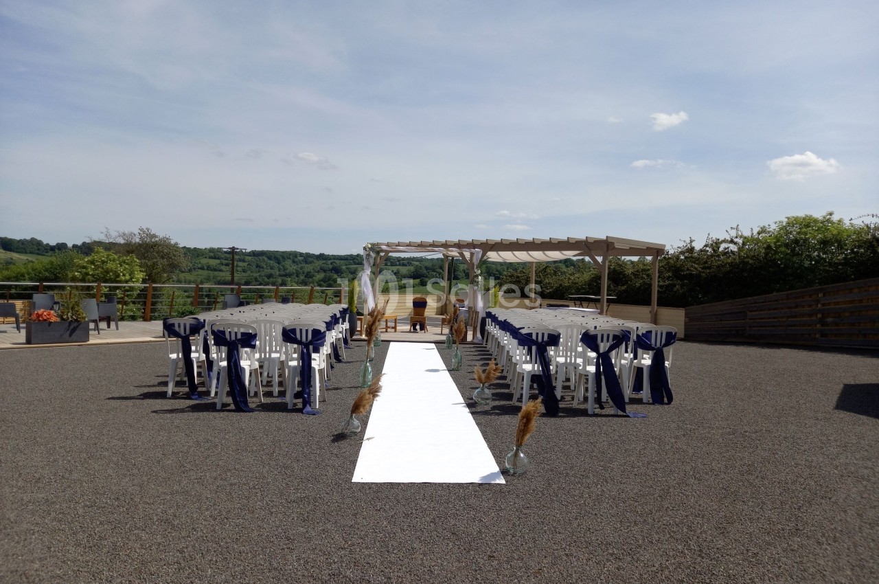 Allée centrale blanche bordée de chaises décorées en extérieur, menant à une pergola avec vue sur la campagne.