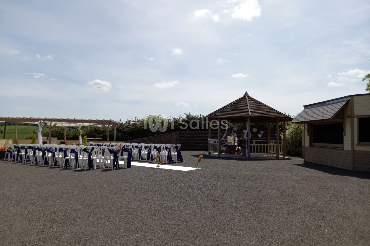 Espace extérieur avec pergola, chaises alignées et kiosque en bois, préparé pour un événement sous un ciel dégagé.
