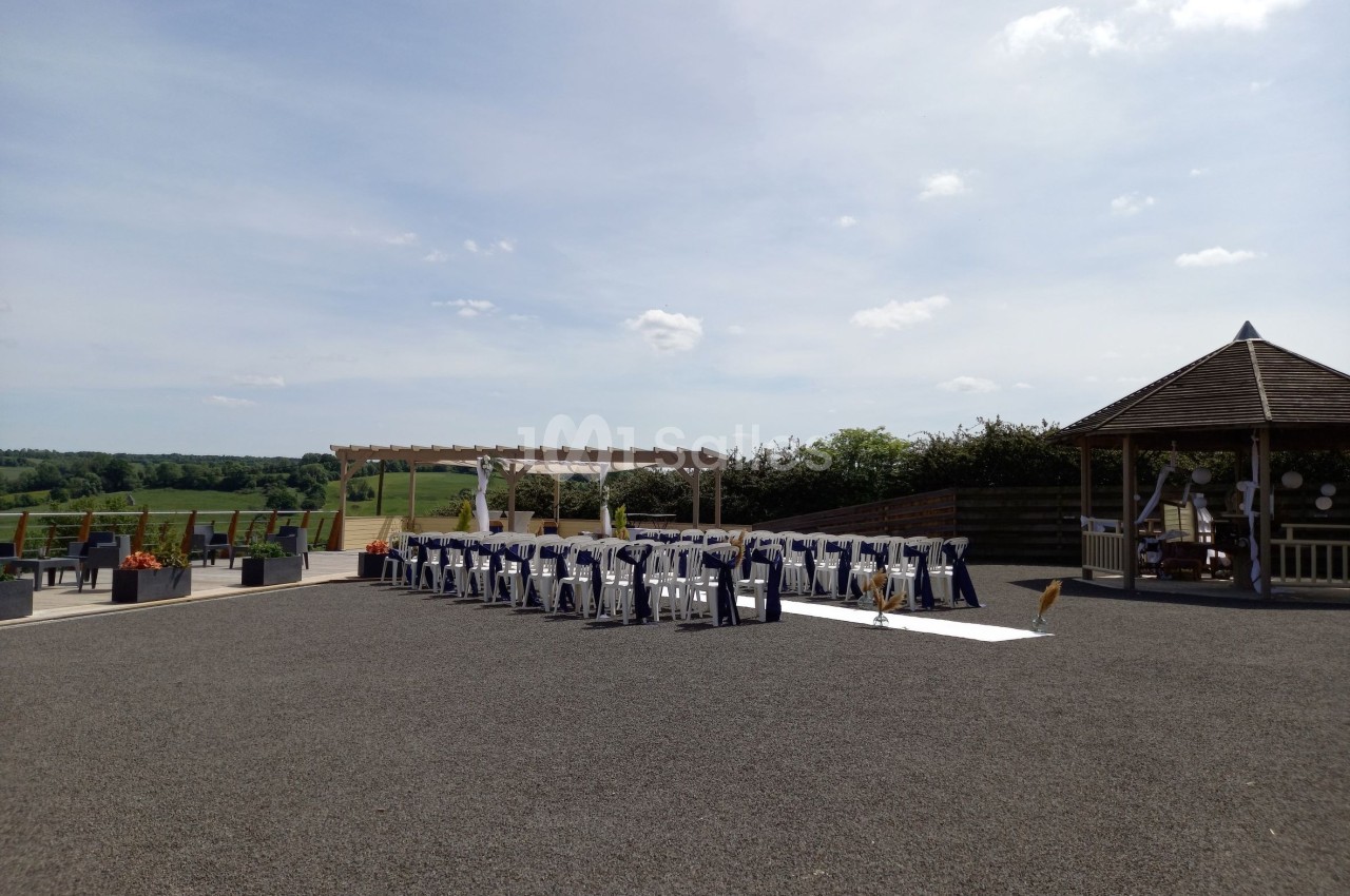 Chaises alignées en extérieur sous une pergola pour une cérémonie, avec vue sur un paysage champêtre et ciel dégagé.
