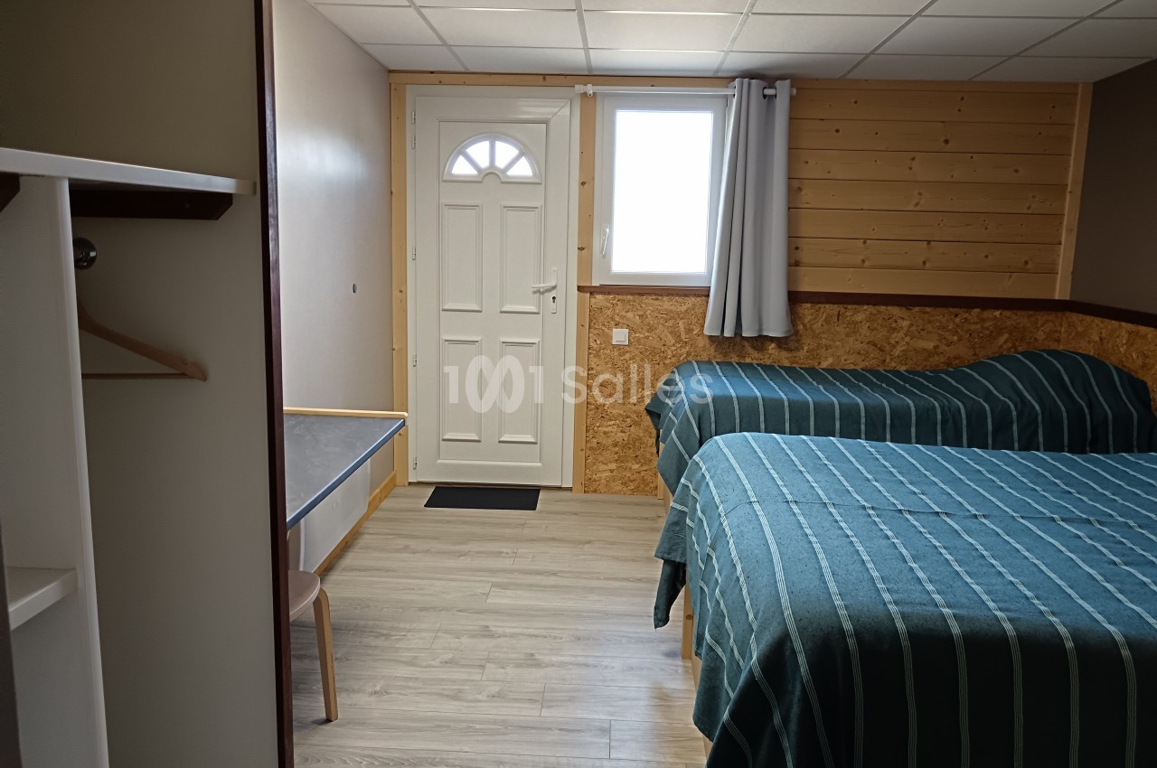 Chambre simple avec deux lits, murs en bois, sol clair, une table, une porte et une fenêtre avec rideau gris.