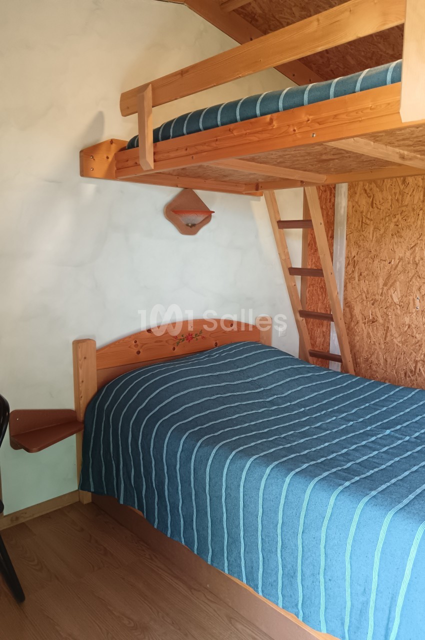 Lit double avec dessus-de-lit bleu, lit superposé en bois au-dessus, échelle et mur en bois dans une chambre simple.