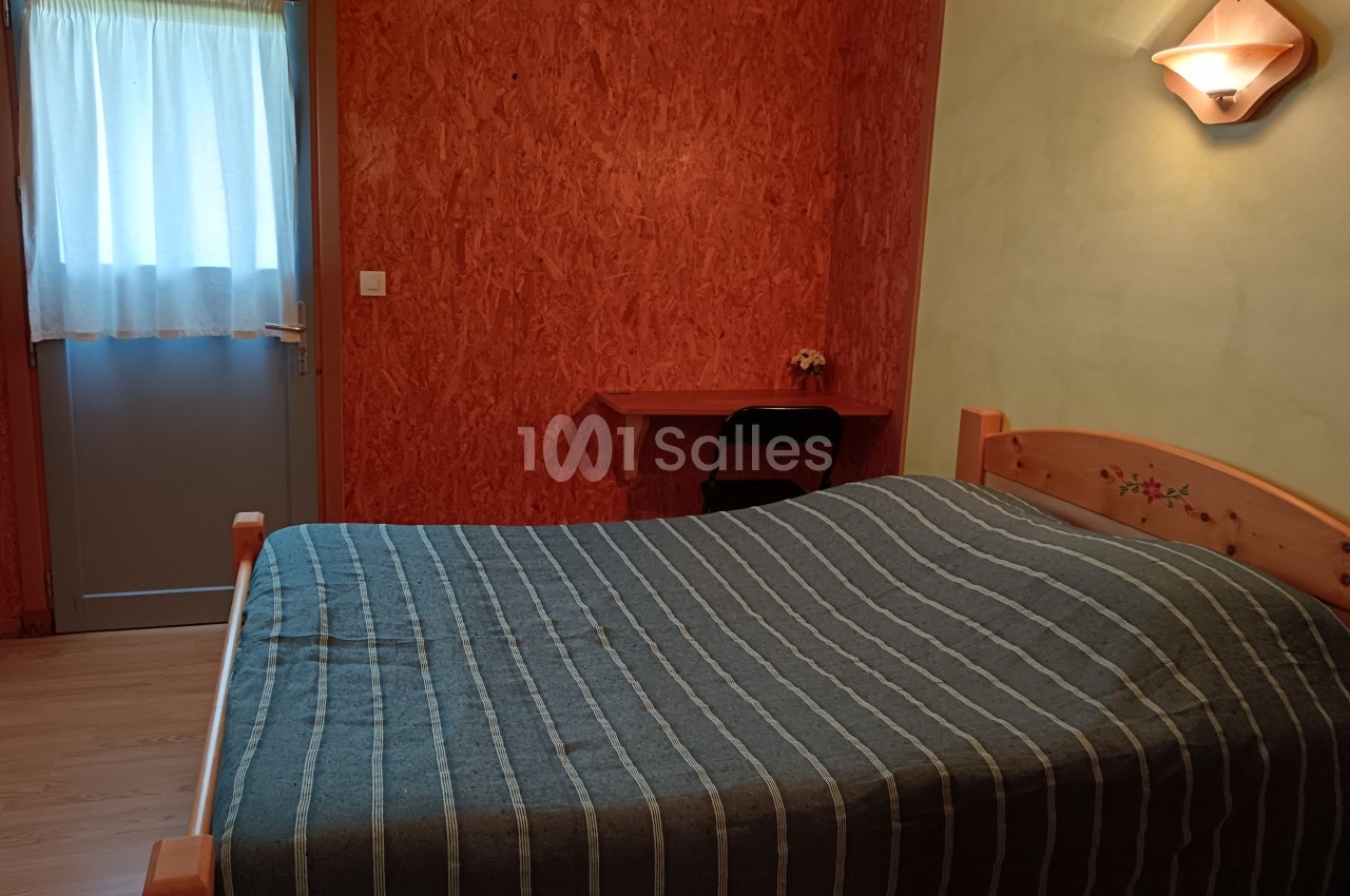 Chambre avec un lit double, une table en bois, une chaise, un mur orange et une porte avec un rideau translucide.
