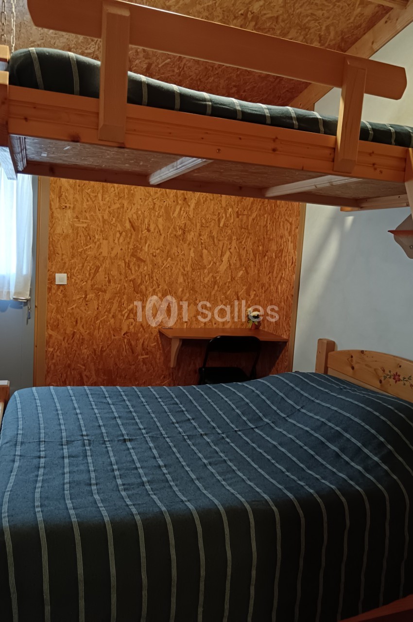 Lit superposé en bois avec une échelle dans une chambre simple, éclairée par une fenêtre avec un bureau en arrière-plan.