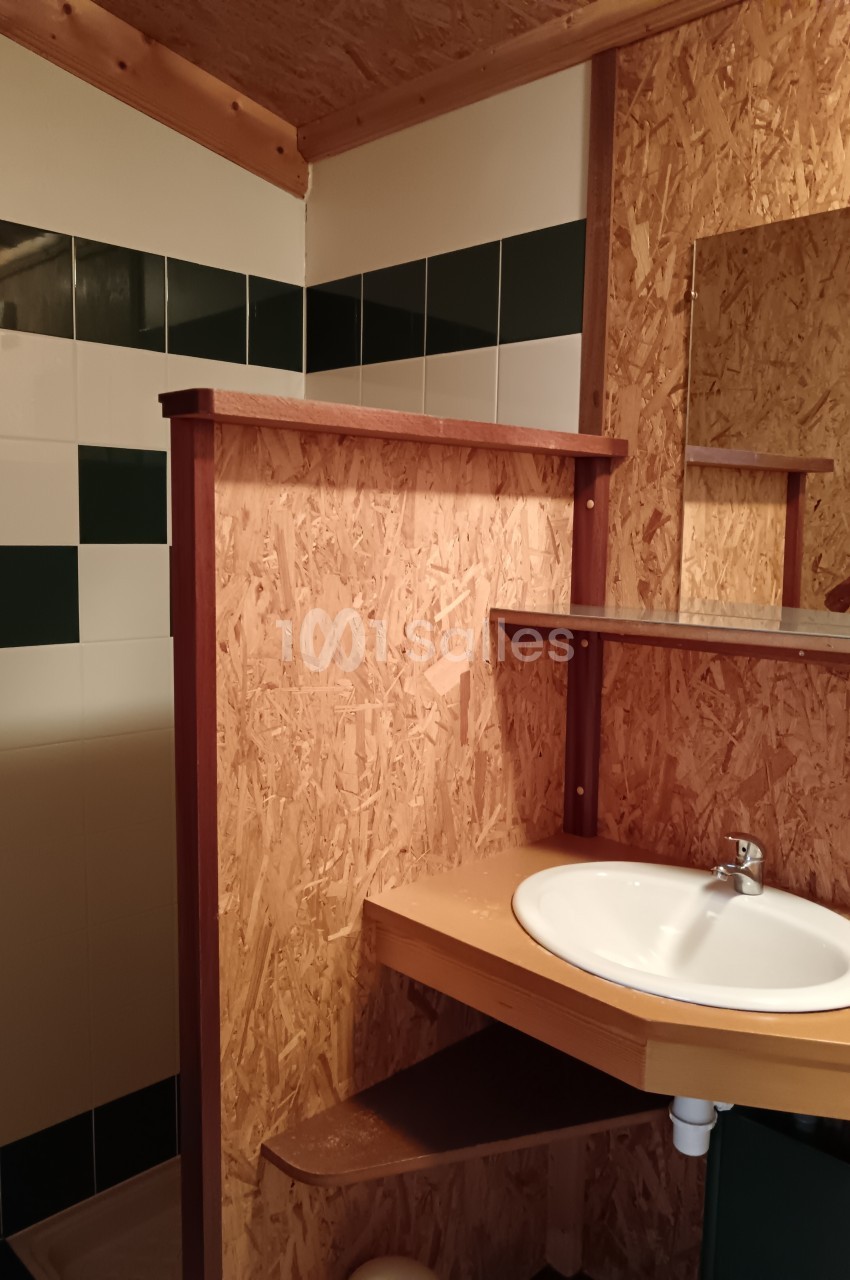 Lavabo avec plan de travail en bois, miroir et cloison en panneaux OSB, dans une salle de bain partiellement carrelée.